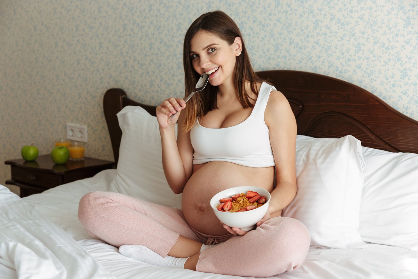 Portrait of a smiling young pregnant woman having healthy breakfast while sitting on bed with a bowl and spoon